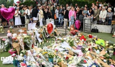 A large crowd of people gather behind a patch of grass in the Annecy park which is covered with bouquets of flowers, balloons and teddy bears.