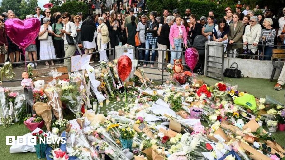 A large crowd of people gather behind a patch of grass in the Annecy park which is covered with bouquets of flowers, balloons and teddy bears.