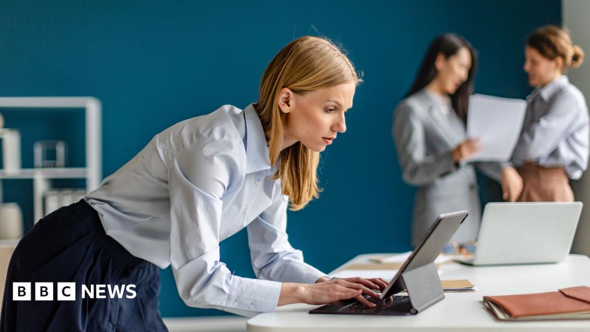 A female office worker wearing a light blue top and dark blue skirt leans over a laptop computer. Two other workers are in the background, slightly out of focus