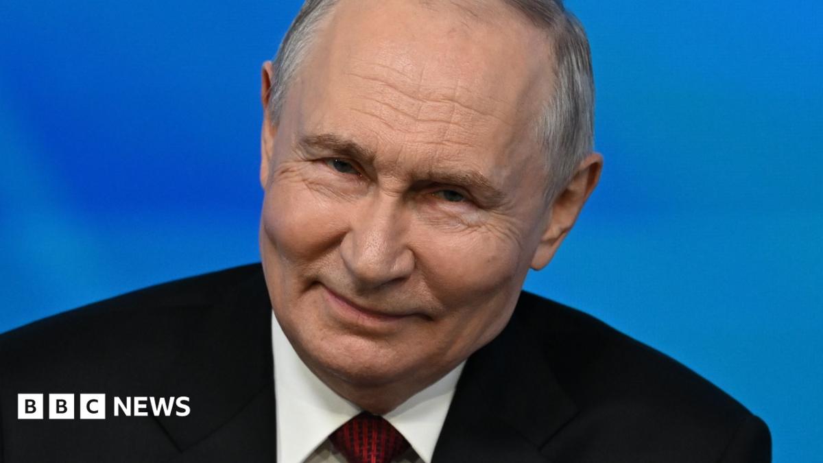Close up of Vladimir Putin while speaking to an audience at the Valdai foreign policy forum. He has short grey hair and is wearing a black suit, white shirt and red tie.
