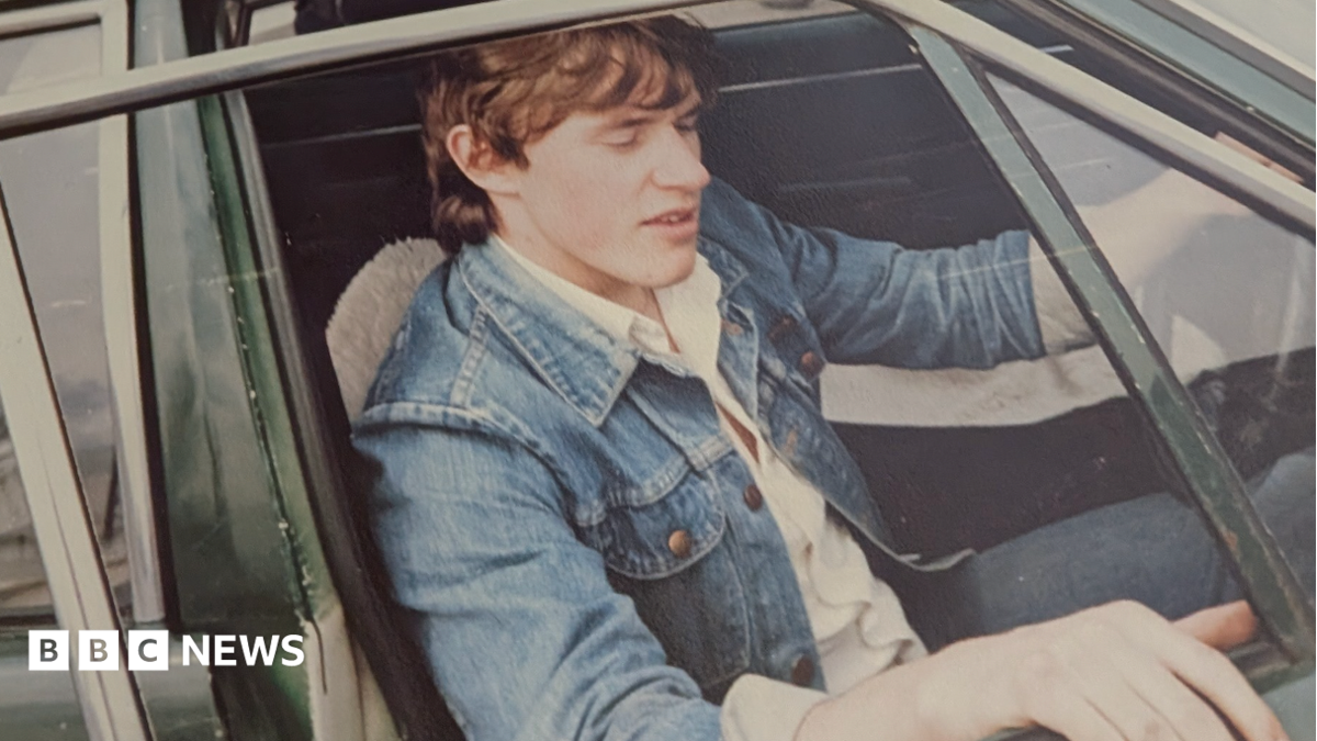 An old photo of a young man with floppy brown hair, sitting in the driver's seat of a car, with the window down and door open. He's wearing a white shirt, jeans and a blue denim jacket.