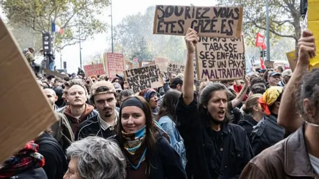 A large number of protesters are shown marching down a street in Paris, shouting and waving placards.