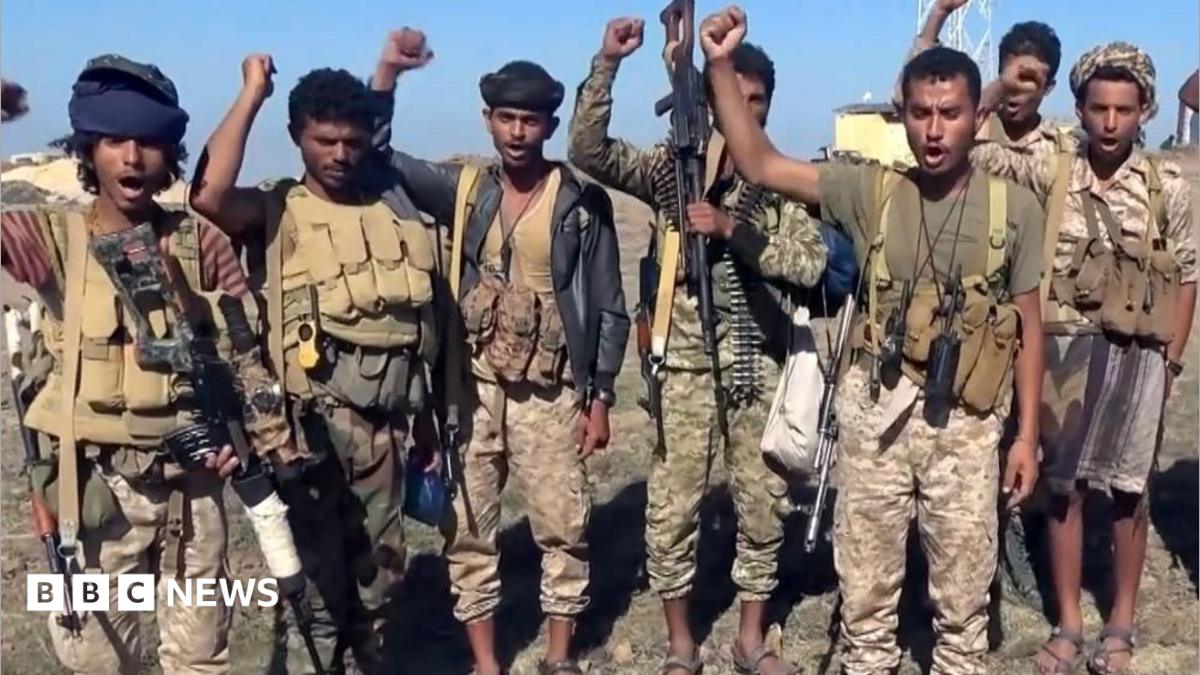 Houthi fighters wearing camouflage uniforms and guns strapped to their shoulders raise fists in the air