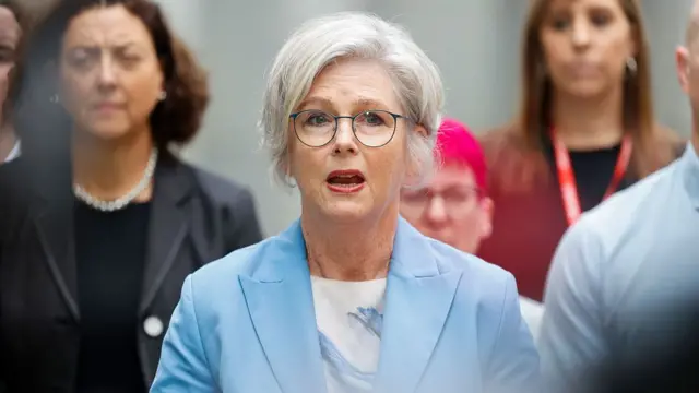 wearing a light blue blazer, white top and round eyeglasses speaks during what appears to be a press conference