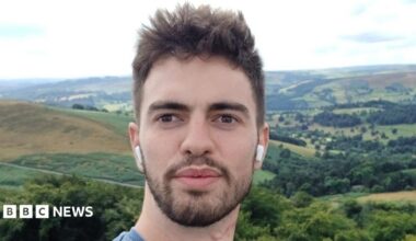 A selfie of William Chapman outdoors, with rolling hills, woodland and patchwork fields stretching across the landscape behind him. He is wearing wireless earbuds and is standing in front of a panoramic countryside view under a bright, cloudy sky.