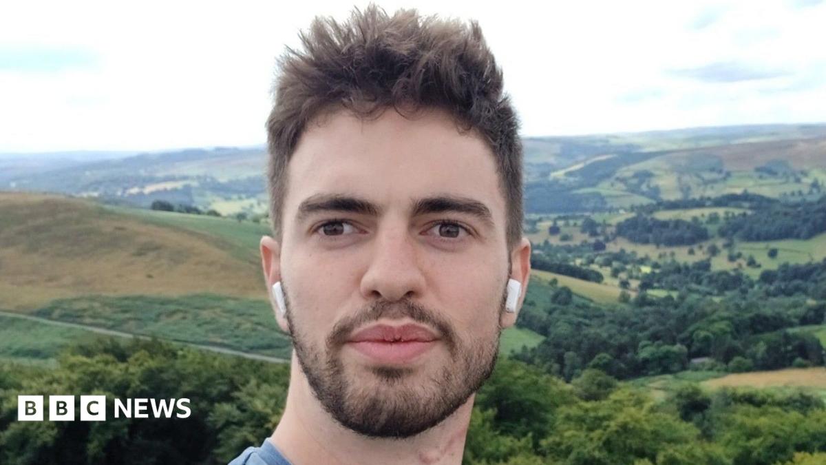 A selfie of William Chapman outdoors, with rolling hills, woodland and patchwork fields stretching across the landscape behind him. He is wearing wireless earbuds and is standing in front of a panoramic countryside view under a bright, cloudy sky.