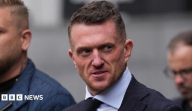 Tommy Robinson stares at the camera as he walks into court, surrounded by a close crowd and security officers. He is wearing a white shirt and black tie. Next to him is a woman with blonde hair and blue eyes, who also stares right at the camera.
