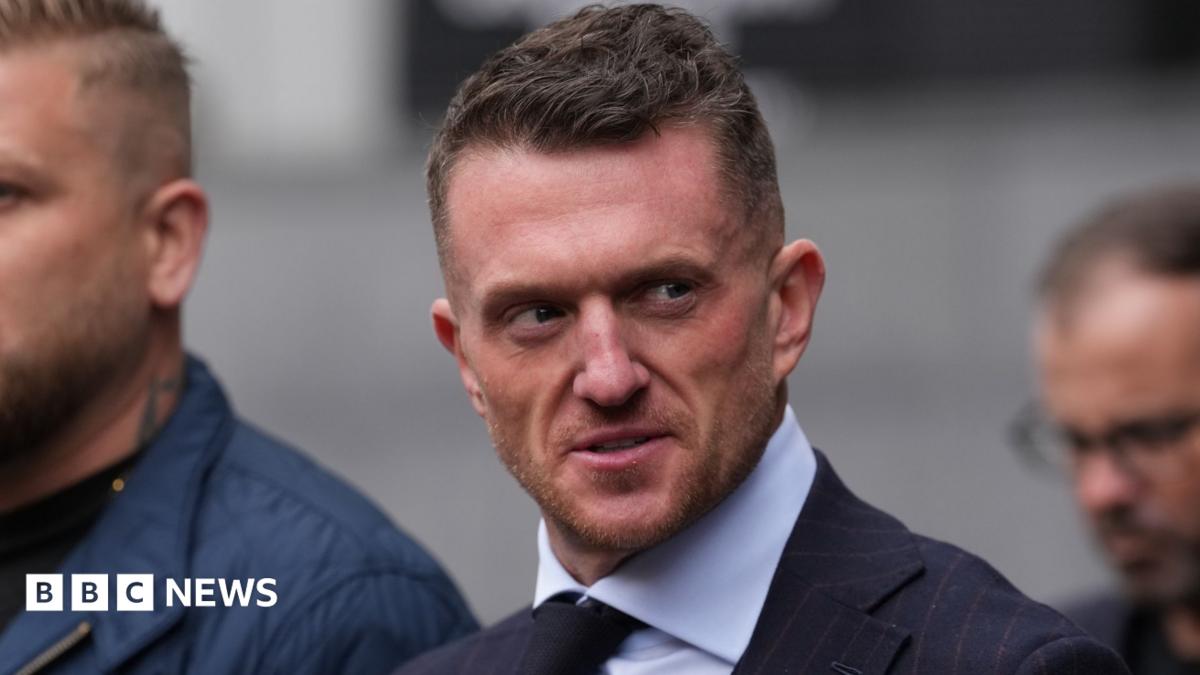 Tommy Robinson stares at the camera as he walks into court, surrounded by a close crowd and security officers. He is wearing a white shirt and black tie. Next to him is a woman with blonde hair and blue eyes, who also stares right at the camera.