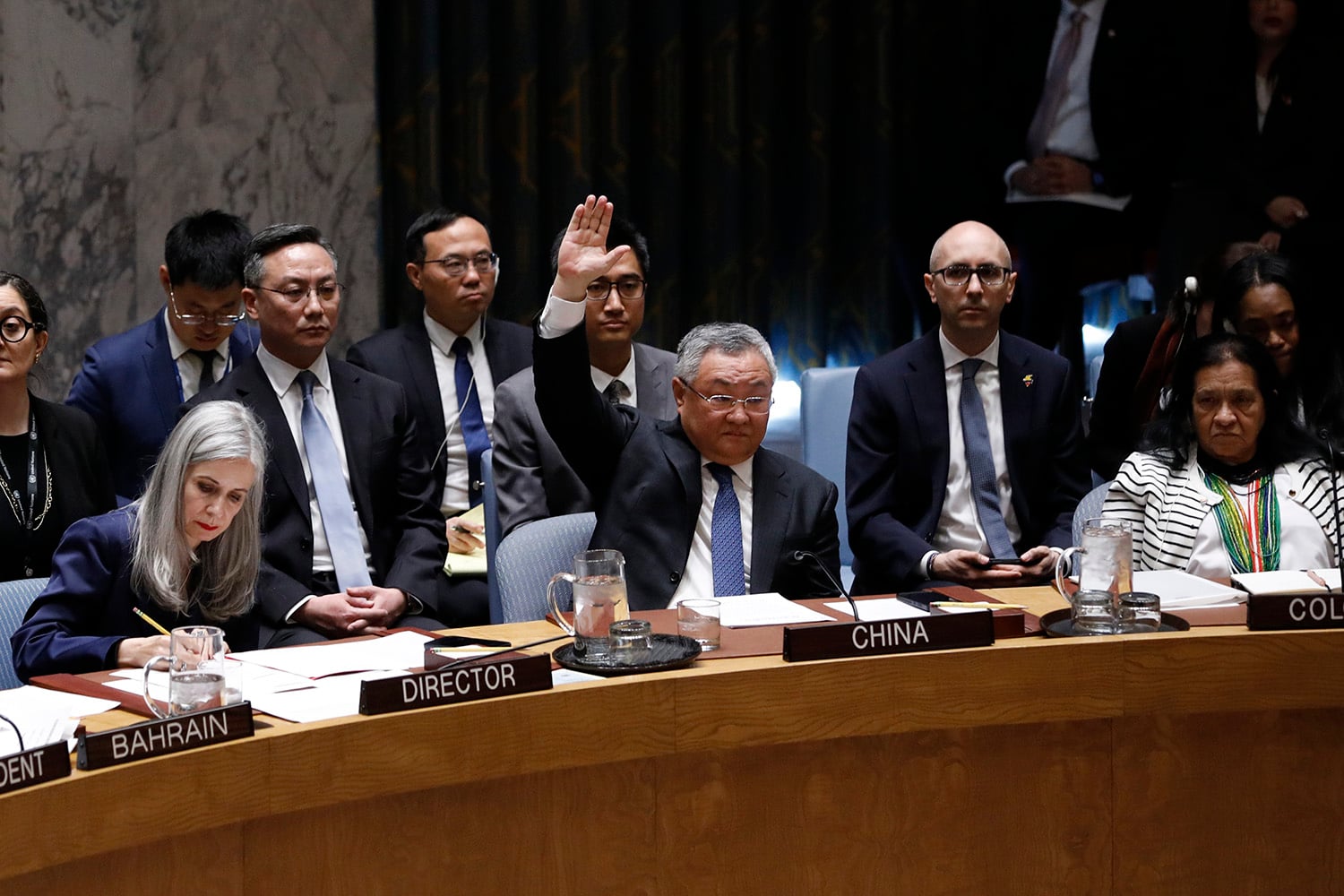 A group of officials sits behind a long wooden dais at an international conference table, with a delegate sitting behind the "CHINA" plaque raising his right hand. The man wears a dark suit and tie and glasses.