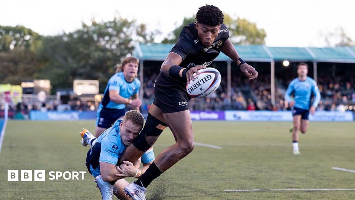 Noah Calouri dives over the tryline with a rugby ball held in his right hand, as Bristol's James Williams wraps around his left leg trying to make a tackle
