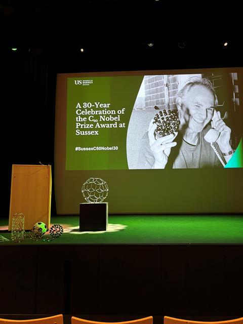 Photo of stage with models of C60, a football and a slide showing Harry Kroto and the words 'A 30-year celebration of the C60 Nobel prize award at Sussex' #SussexC60Nobel30