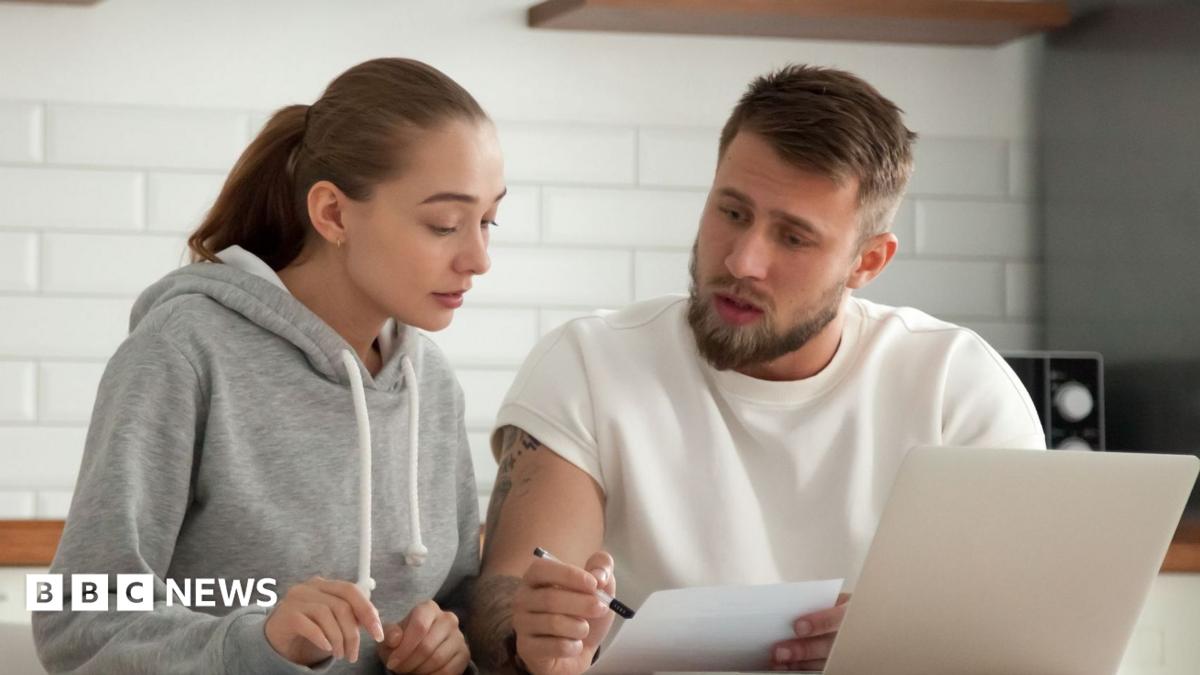 Couple look at a bill in their kitchen with a laptop in front of them.