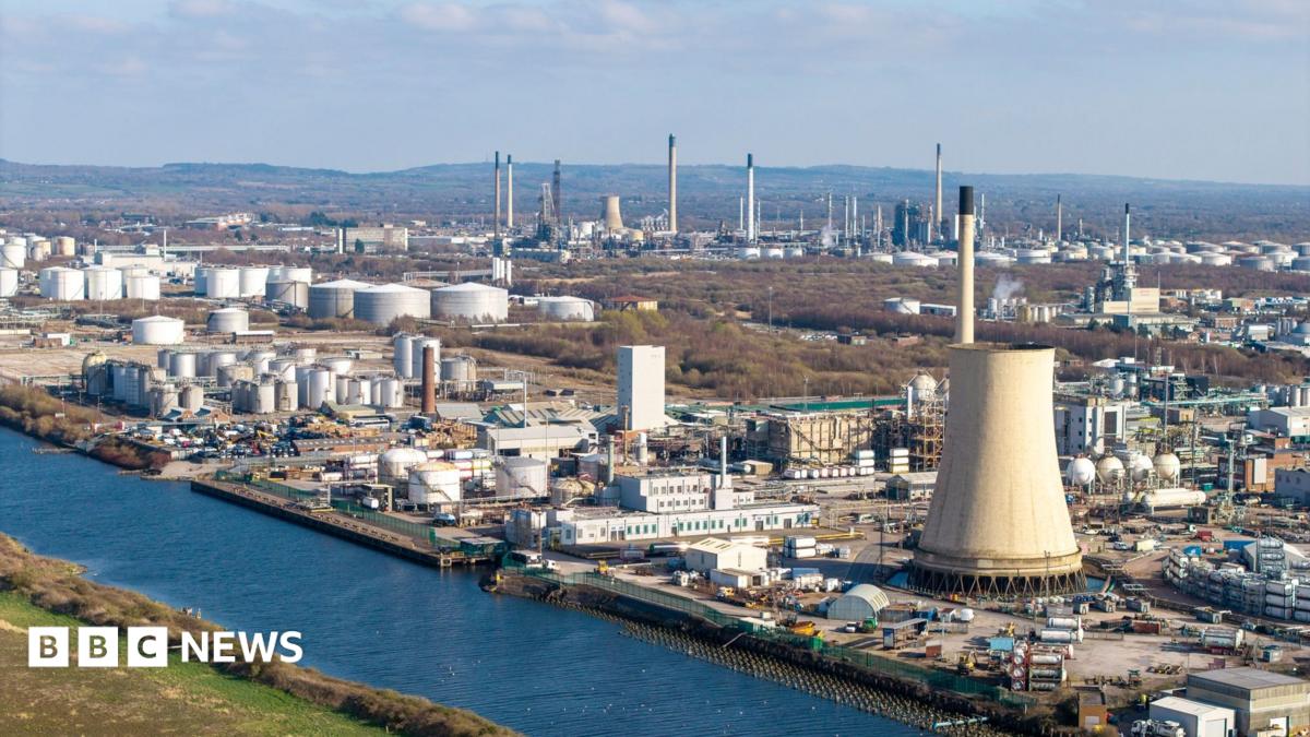The Manchester Shipping Canal can be seen running across the image, on the left bank is grass and on the right a large industrial complex stretching into the distance. There are rolling hills outlined in the background and a blue sky with some white clouds.