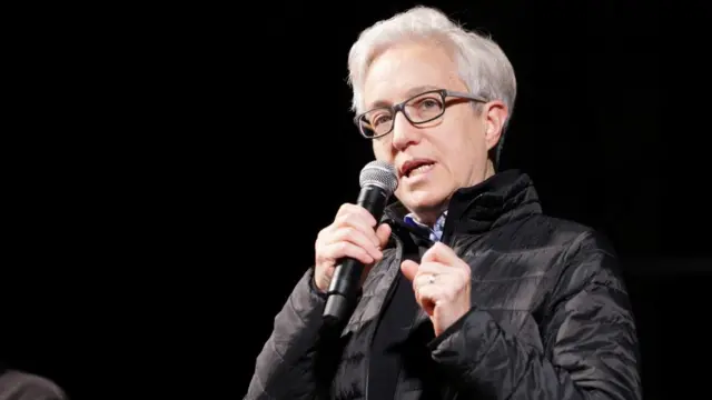 Oregon Governor Tina Kotek speaking at a candlelight vigil with a black puffer jacket and a microphone