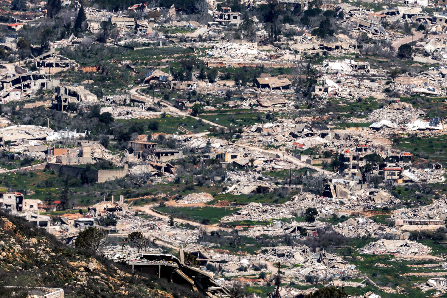An elevated wide view shows a stretch of a vast, heavily damaged area covered in rubble, flattened low-rise buildings, and scattered debris.