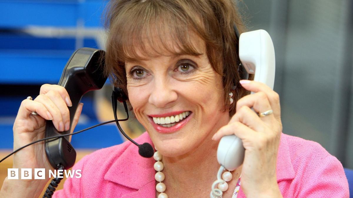 Close-up of Dame Esther Rantzen in an office wearing a headset and a pink jacket, holding two telephone handsets, one black and one white. A blue-toned workspace is visible in the blurred background. She is wearing a white beaded necklace is worn around the neck. She is smiling