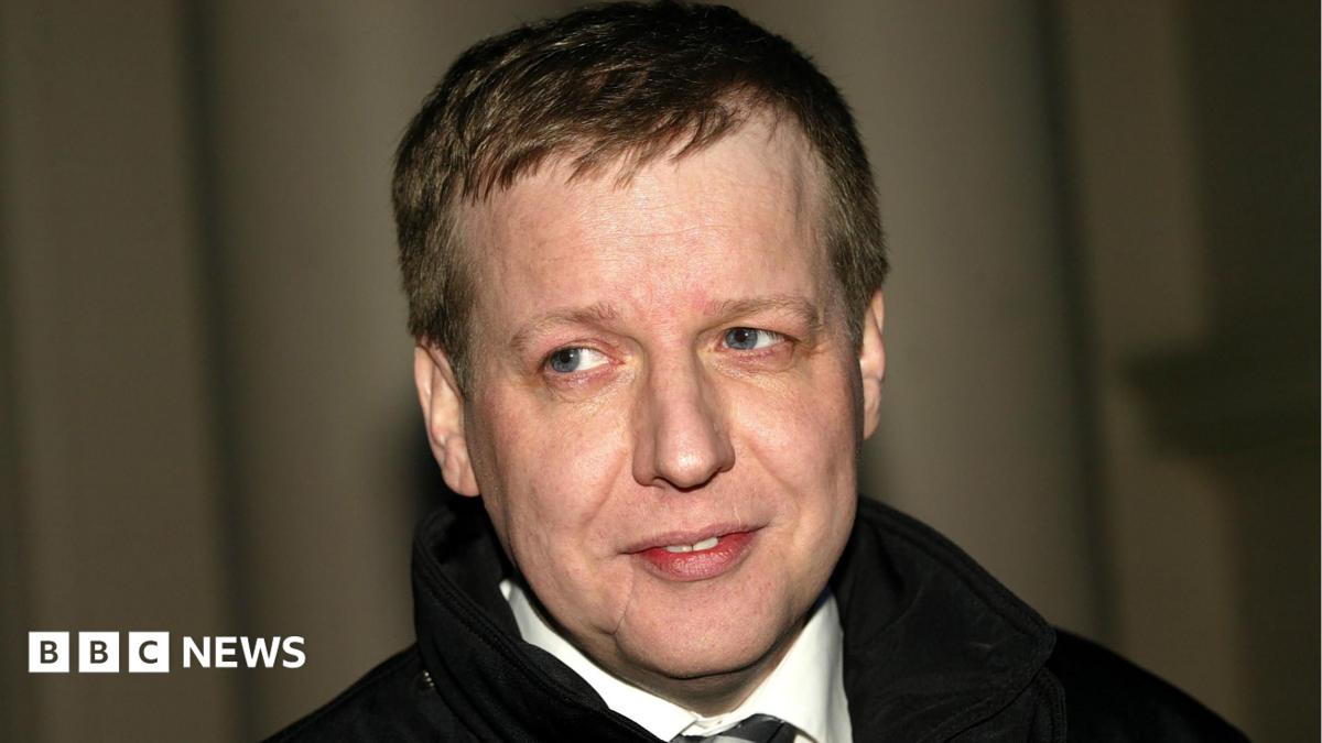 Paul Ferris, a pale man with a scarred face, is wearing a dark coat, white short and dark tie. He is smiling and looks off camera against a curtain backdrop.
