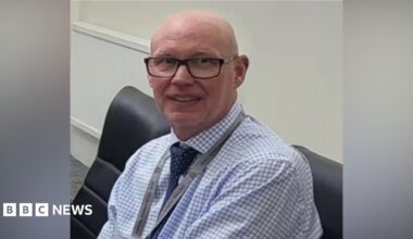 MI5 chief Sir Ken McCallum has short dark hair, dark glasses and is wearing a dark suit with a green tie.