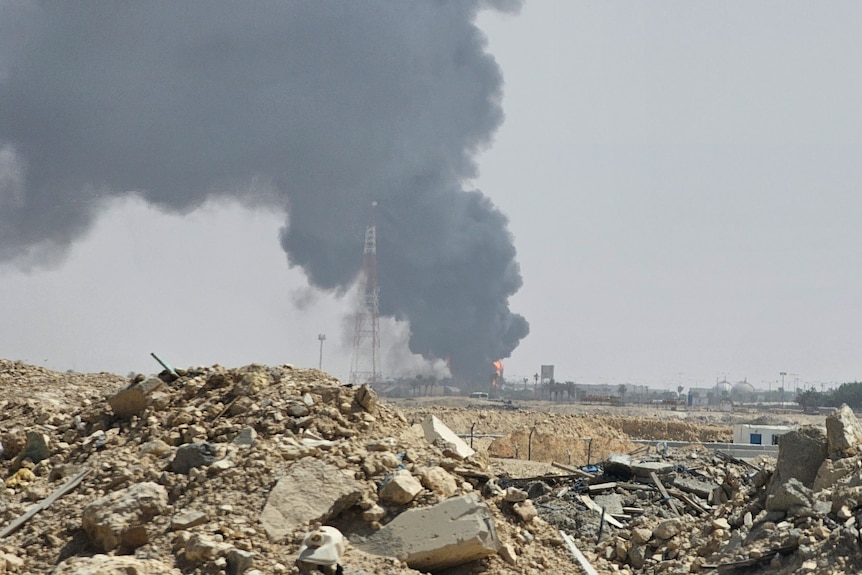 A large cloud of black smoke and flames rise out of an oil facility with rubble in the foreground.