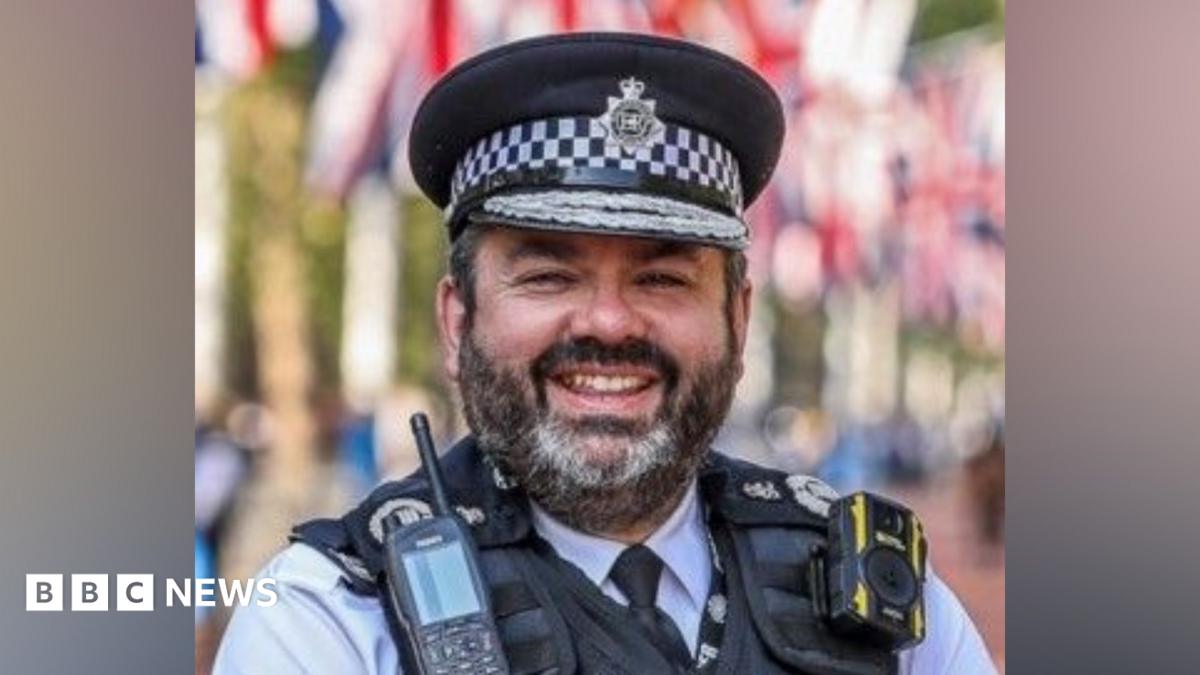 A man in a Metropolitan Police uniform and peaked cap stands outdoors, wearing a radio and body-worn camera on a protective vest. Red, white and blue flags are visible blurred in the background.
