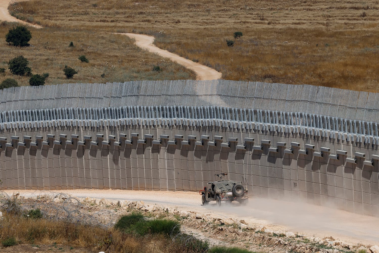 A military vehicle drives away along a dirt road adjacent to a tall concrete border wall topped with high wire fencing. The wall is at least five times taller than the vehicle. A dry landscape covered in grasses and small shrubs is visible beyond the fence.