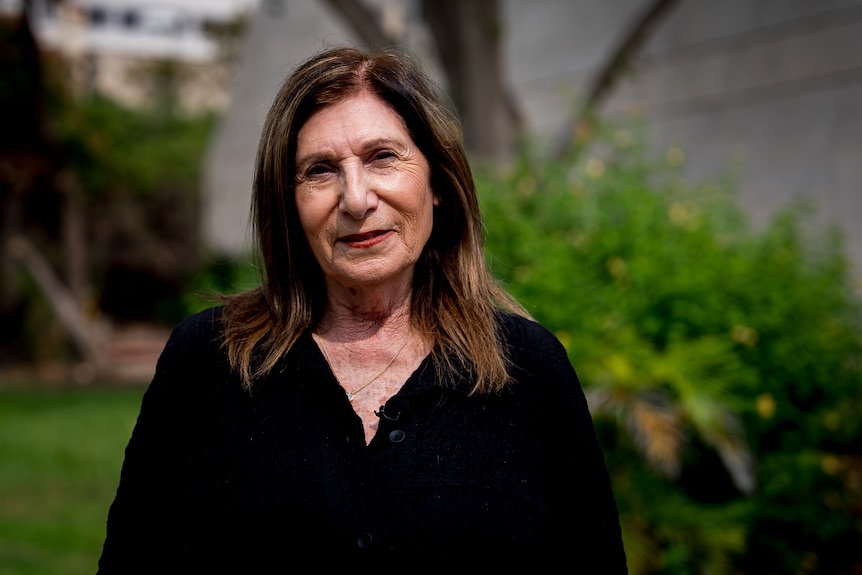 A middle aged woman with dark hair smiles and stands in a garden.