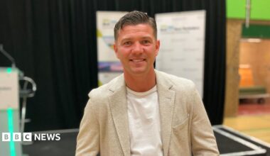 Luke Campbell standing by a stage on election night. He has short dark hair and is wearing a cream jacket and white T-shirt