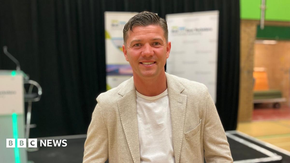 Luke Campbell standing by a stage on election night. He has short dark hair and is wearing a cream jacket and white T-shirt