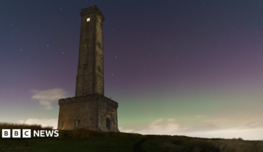 The Peel Tower stands on a hillside beneath a star-filled night sky, illuminated by a green and purple aurora glow on the horizon. Low clouds drift across the landscape below the historic stone monument.