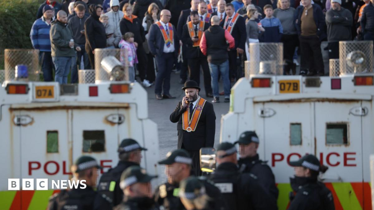 Image of two police rovers, white with the word police written in red. At the bottom of the van is a red and yellow striped pattern. Metal guards cover the sirens of the vehicles. Images of police wearing black caps are at the forefront of the image. Directly behind them is a man with a black hat, orange sash and dark suit on the phone with someone. Other people in similar outfits are stood behind him.