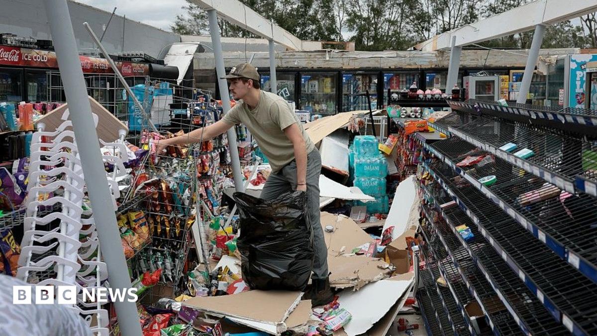 A man puts rubbish in a bag with debris strewn all around a store with the roof blown off