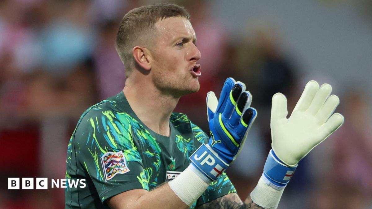 Jordan Pickford is wearing the England goalkeeper strip. He has short brown hair and is clapping his keeper gloves together.