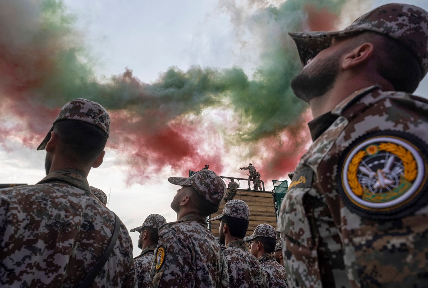 A low-angle view of personnel in camouflage uniforms looking up toward the sky as red and green smoke billows above them. The servicemen also wear camouflage brimmed hats, obscuring their faces.