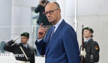 German Chancellor Friedrich Merz in a blue suit with hand raised, two military officers stand behind him, blurred.