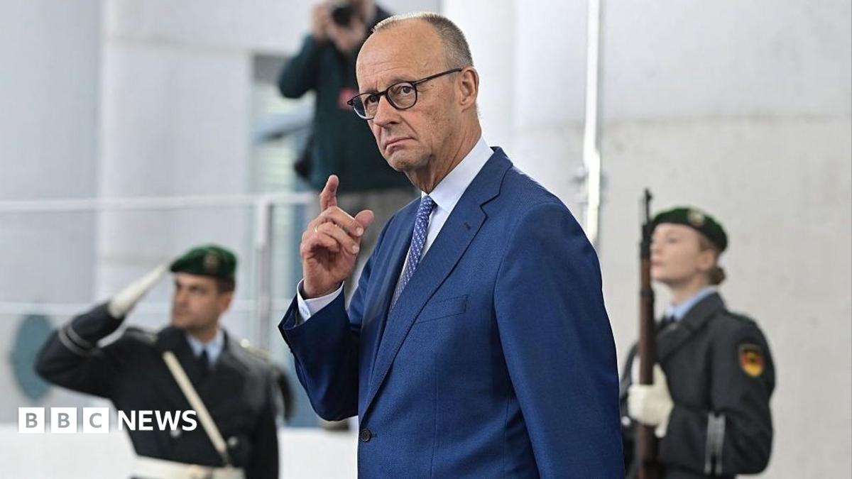 German Chancellor Friedrich Merz in a blue suit with hand raised, two military officers stand behind him, blurred.