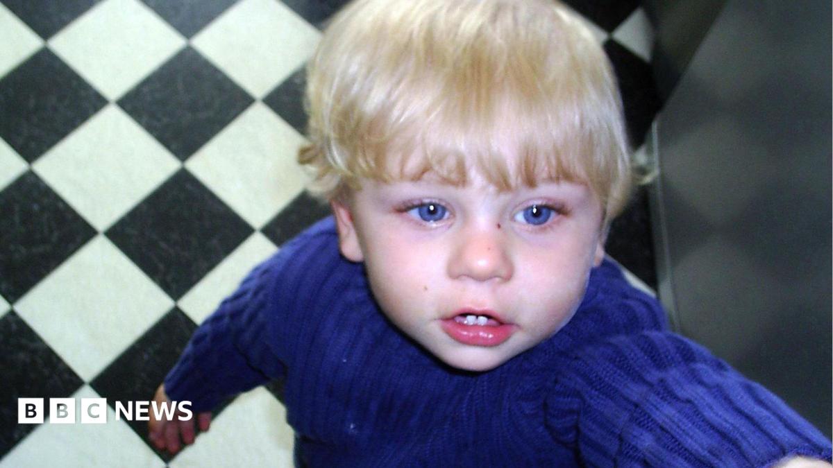 A toddler boy with blonde hair and blue eyes seen standing with photo taken from above
