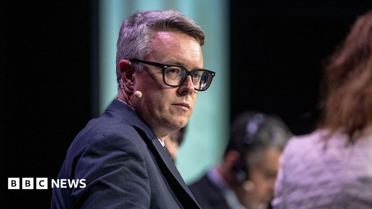 Energy Minister Martin McCluskey, a white man with glasses pictured on stage at an event, with an earpiece and microphone wearing a navy suit.