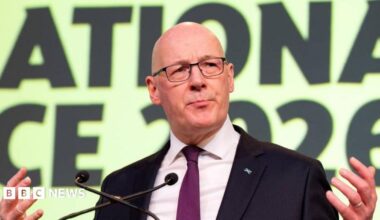 John Swinney, a bald man with glasses wearing a black suit and purple tie with a saltire pin on it. He has his hands outstretched beside him and two microphones in front of him. He is mid speech. Behind him is a yellow sign with black writing.
