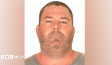A police mugshot of a man wearing a grey t-shirt. He has very close-cropped short brown hair and stubble. He has a chubby face and neck of roughly the same width and appears to be sunburnt.