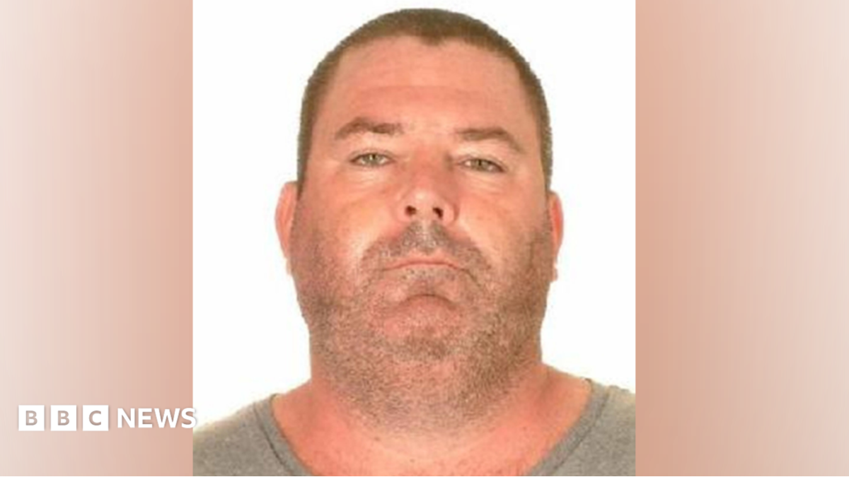 A police mugshot of a man wearing a grey t-shirt. He has very close-cropped short brown hair and stubble. He has a chubby face and neck of roughly the same width and appears to be sunburnt.