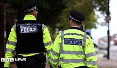 Two Met Police officers face away from the camera as they walk down a street in London. Both are wearing hats and hi-viz jackets. One on the left wheres a black vest over his jacket.