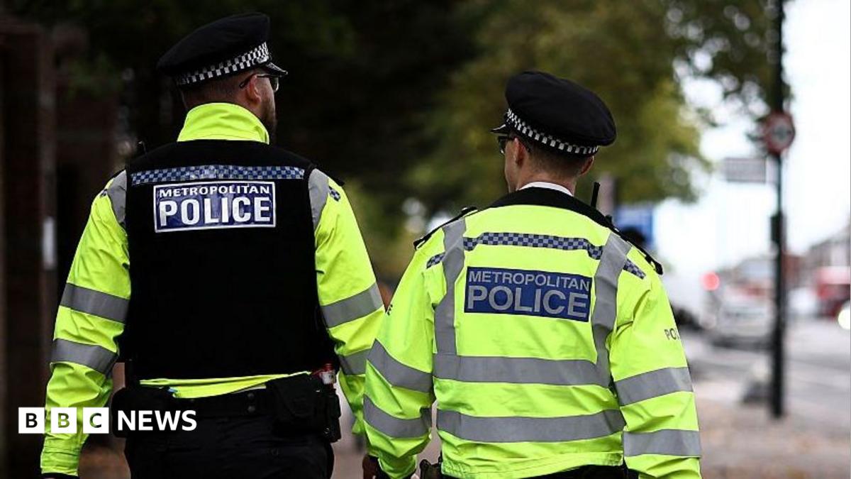 Two Met Police officers face away from the camera as they walk down a street in London. Both are wearing hats and hi-viz jackets. One on the left wheres a black vest over his jacket.
