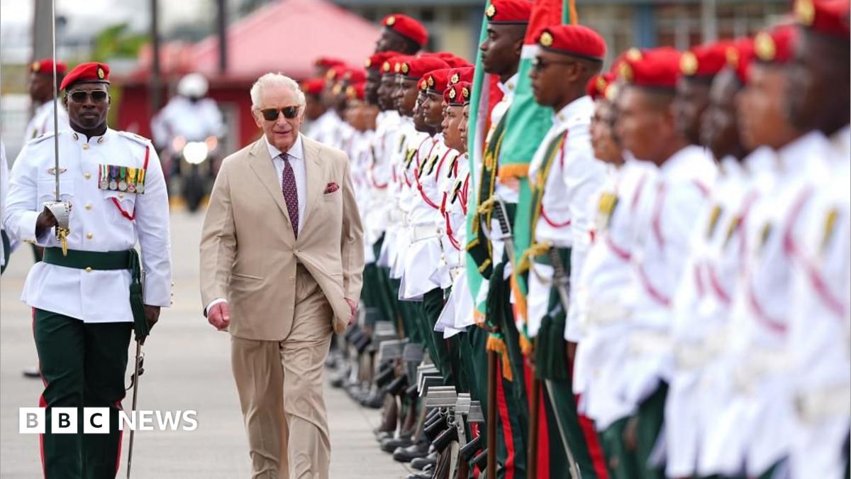 King Charles III, in a light brown suit, observes a guard of honour. He walks along a line of members of the Guyana Defence Force, who wear white shorts with military honours on them, and green trousers.