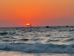A sailboat cruises against an orange sky at sunset as waves crash on the beach.
