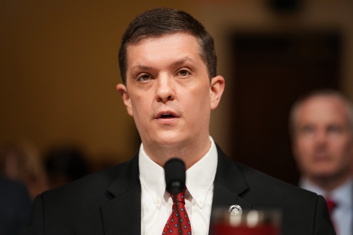A man wearing a suit and tie speaks into a microphone.