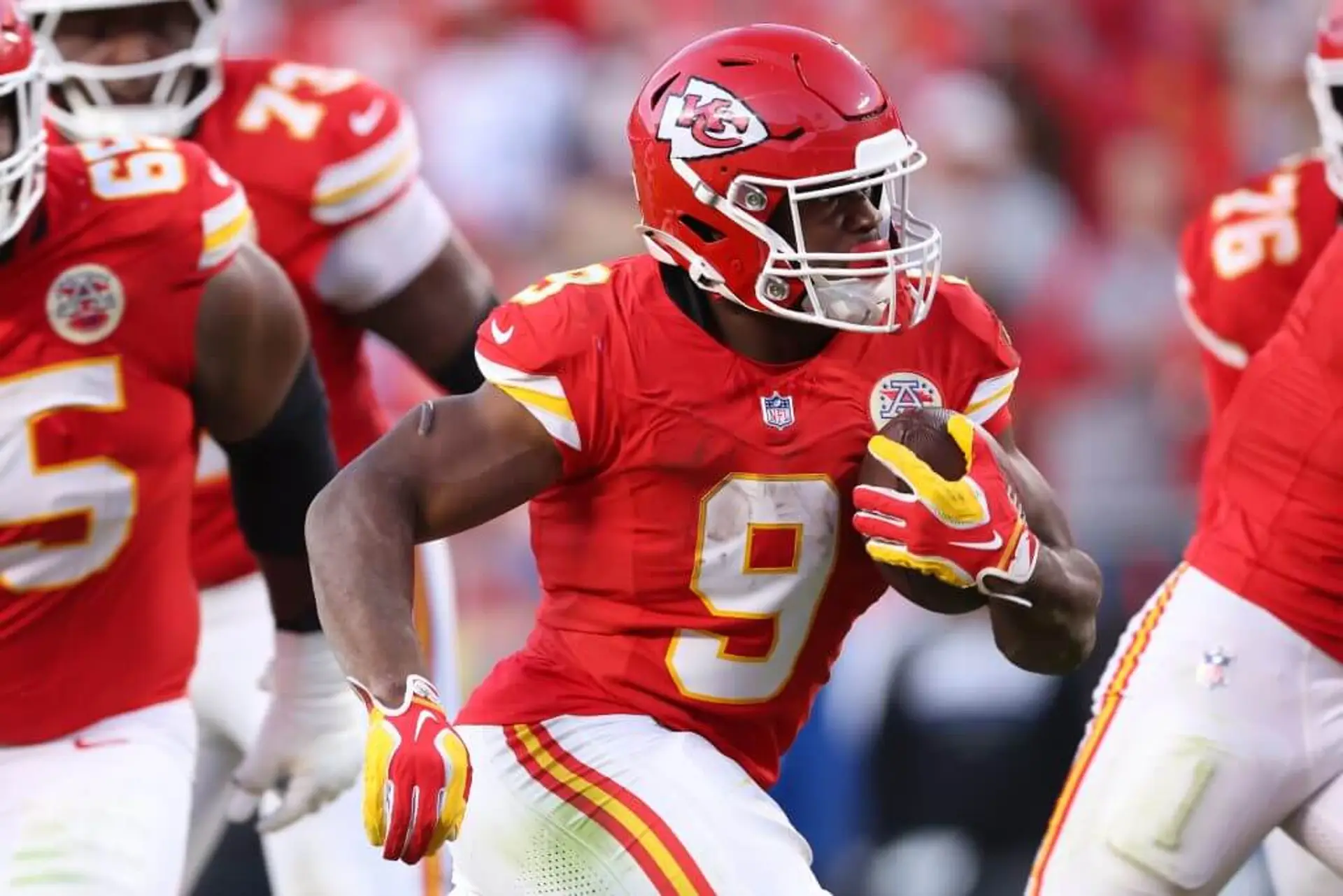 A football player in a red jersey and helmet runs with the ball in his left arm.