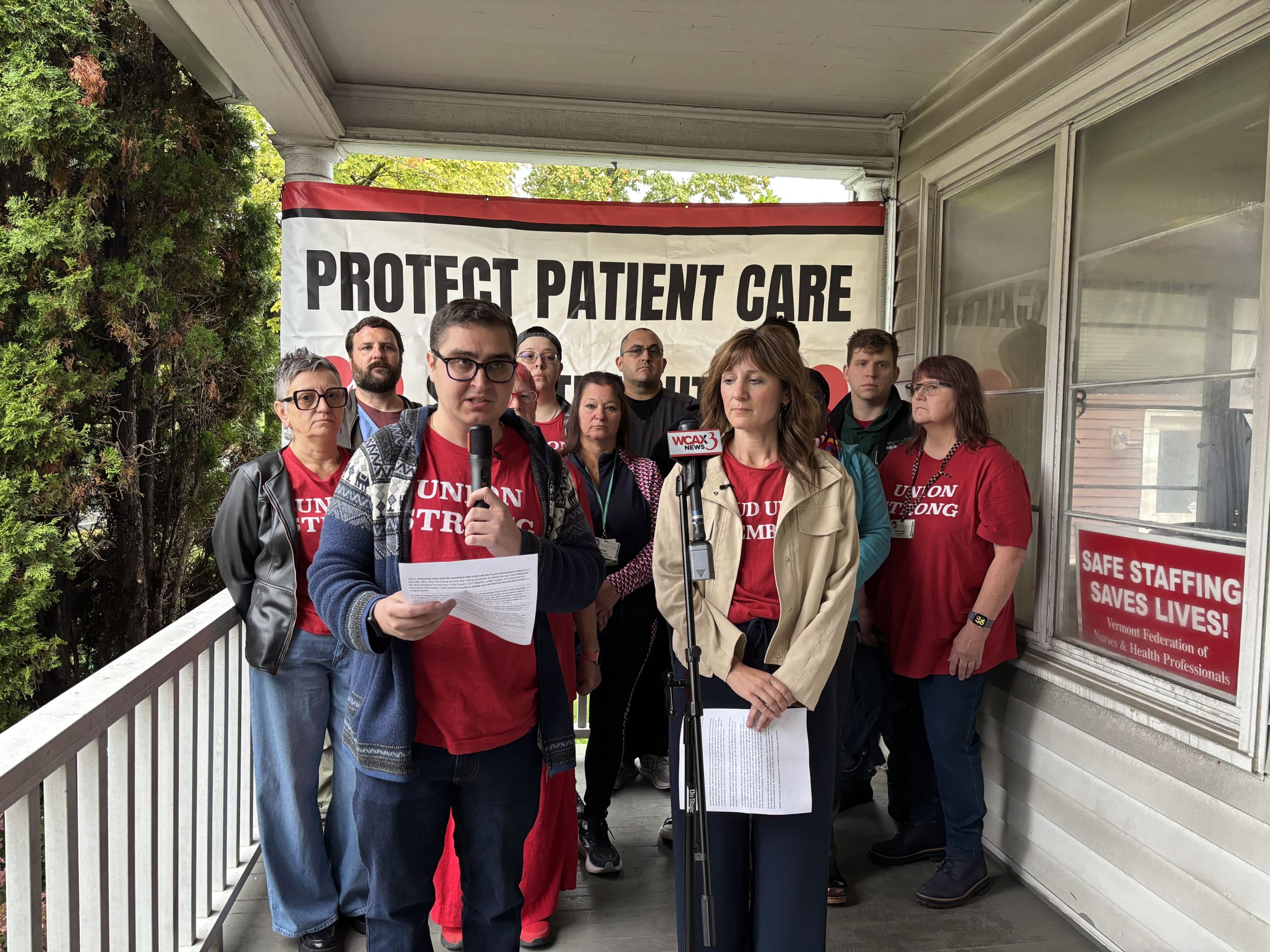 A group of nurses in “Union Strong” t-shirts stands on a porch behind two speakers holding microphones and papers in front of a “Protect Patient Care” banner.