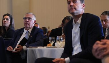 On the left, a man in his 60s with short grey hair wearing a suit leans back in a chair. On the right, a middle-aged man with short brown hair wearing a suit leans forward. The two men are sitting at a hotel conference room table.