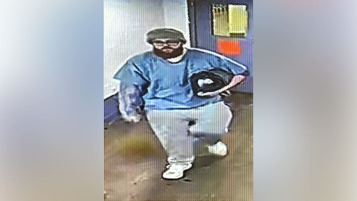 Surveillance image of Jacob Allen Morley wearing a blue shirt, light-colored pants and a dark cap while walking through a prison facility.