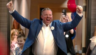 A light-skinned man in a blue blazer and white button-up shirt pumps his fists in celebration in a room full of people.